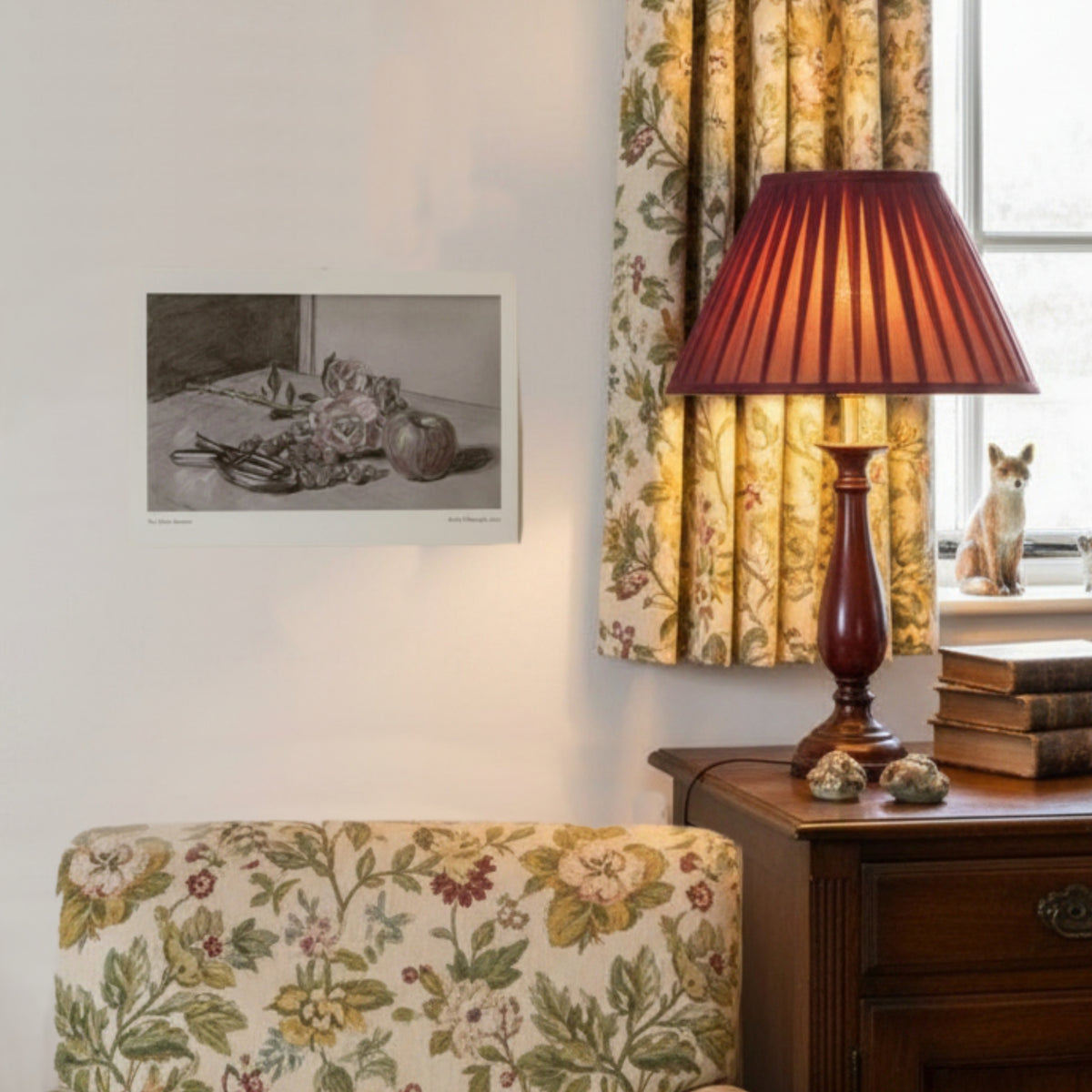An art print showing flowers and fruit on a white wall with chintz curtains and chairs and a lamp