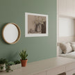 A sepia-toned still life art print depicting a vase of flowers and a bowl of eggs, hanging on a green wall in a styled room with pot plants, mirror and window