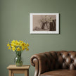 A sepia-toned still life art print depicting a vase of flowers and a bowl of eggs, hanging on a green wall in a styled room with a leather armchair and vase of daffodils