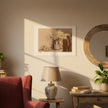 A sepia-toned still life art print depicting a vase of flowers and a bowl of eggs, hanging on a beige wall in a styled room with a red armchair, table lamp, books, and a round wooden mirror.
