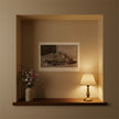 An art print showing flowers and fruit on a cream wall in an alcove with flowers and a lamp