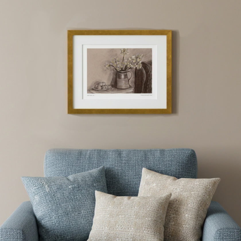 A framed still life art print featuring a pitcher with flowers, displayed on a beige wall above a blue armchair with textured pillows.