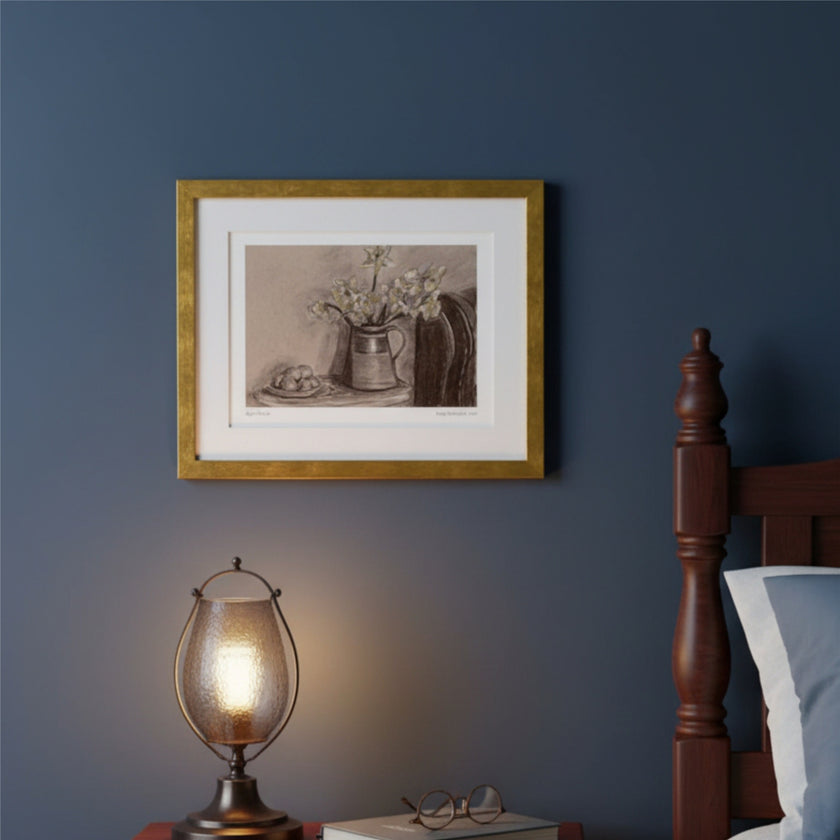 A framed sepia-toned still life artwork with a vase of flowers, displayed on a dark blue wall above a bedside table with a lit lamp and part of a wooden bed visible.