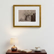A framed still life art print featuring a pitcher with flowers and a small plate, displayed on a white wall above a wooden table with a lit lamp, open book, and teacup.