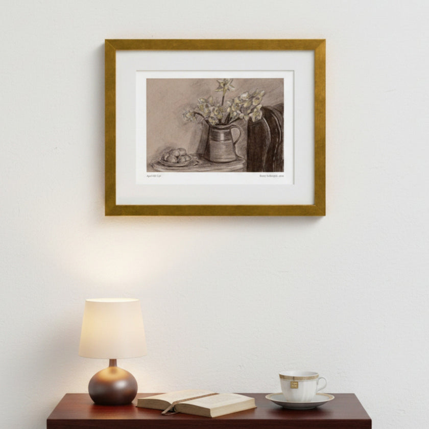 A framed still life art print featuring a pitcher with flowers and a small plate, displayed on a white wall above a wooden table with a lit lamp, open book, and teacup.