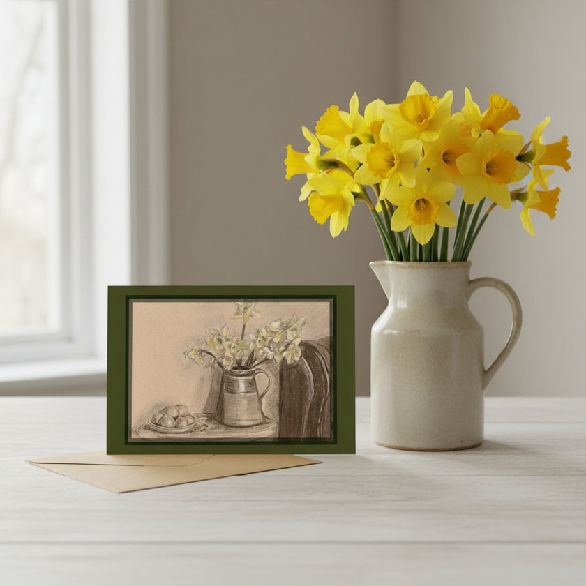 A greetings card depicting a still life of daffodils in a jug, styled on a light wooden table next to a ceramic pitcher filled with yellow daffodils. The print has a dark green frame and is shown with a beige envelope.