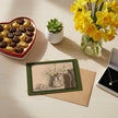 A greetings card showing a jug of daffodils next to a vase of daffodils, a pot plant, chocolates and a necklace in a box.