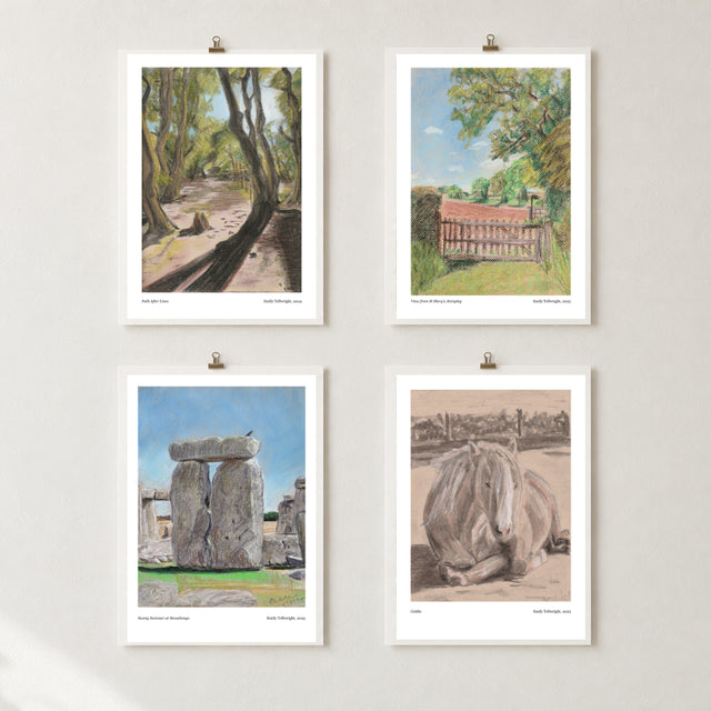 Four framed art prints hanging on a light gray wall with small clips. The prints show landscape and animal scenes: a tree-lined path, a bench in a park, Stonehenge, and a resting horse.
