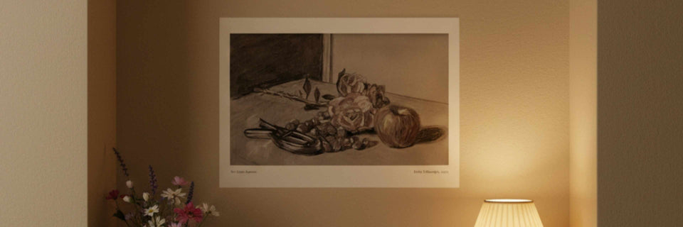 A framed monochrome still life art print hanging on a warm beige wall, with a lit lamp and flowers visible in the room setting.