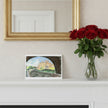 A greetings card depicting Biddulph Grange seen through a stone archway, styled on a white mantelpiece next to a vase of red roses and beneath a gold mirror.