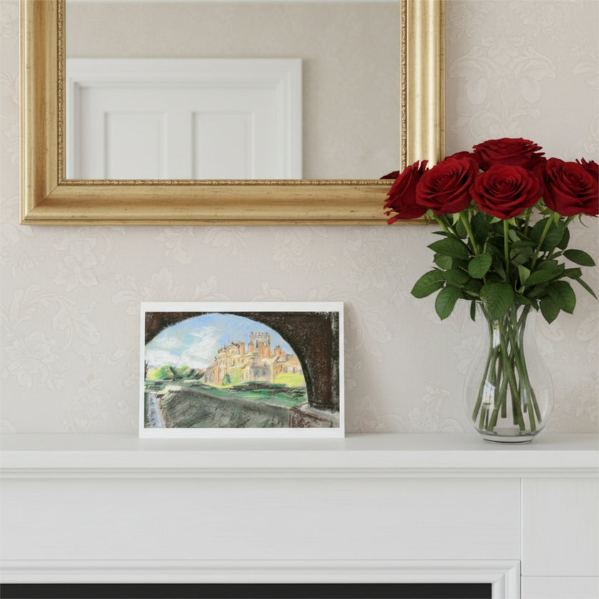 A greetings card depicting Biddulph Grange seen through a stone archway, styled on a white mantelpiece next to a vase of red roses and beneath a gold mirror.