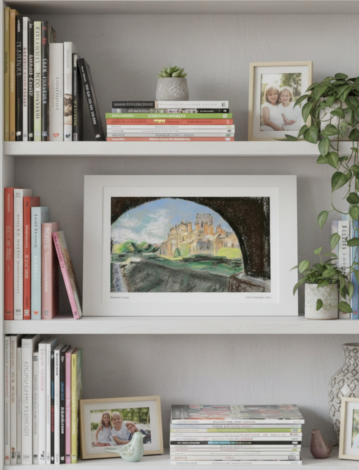 A mounted art print depicting Biddulph Grange through an archway, styled on a white bookshelf alongside books, small potted plants, and framed photos.