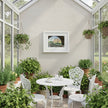 A bright sunroom or conservatory corner with a small white round table and three matching white chairs arranged around a central white pitcher and cup. The room is filled with potted green plants and hanging greenery, with a framed landscape print on the wall.