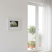 A small framed art print with a white frame hangs on a white wall next to a white-framed glass door looking out onto a balcony with potted plants.