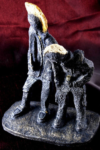 A dark ceramic figurine of two stylised  punk figures on a rectangular base, photographed against a dark red fabric backdrop.