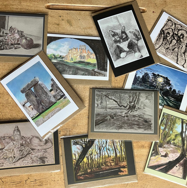 A collection of art greetings cards  arranged on a wooden table. The prints feature monochrome charcoal drawings, landscape scenes, and figures, with some showing visible wood frames.