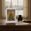 A vertical greeting card featuring an illustration of a decorated Christmas tree, displayed on a wooden desk next to a black ink bottle, gold pen, and envelope. The card is set against a real-world background with a window showing a snowy outdoor scene.
