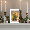 A greetings card a decorated Christmas tree, displayed on a mantelpiece flanked by lit white candles in gold holders, greenery, and white ornaments.