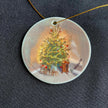 A round ceramic ornament showing a christmas tree with a hole threaded with a gold cord all on a black background