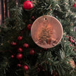 a round ceramic ornament showing a christmas tree hanging on decorated fir foliage