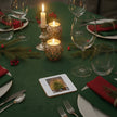 A festive Christmas table setting with a green tablecloth, lit candle holders, wine glasses, silverware, red napkins, evergreen branches, and a small square photo print on a coaster in the foreground.