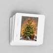 A stack of square white coasters with rounded corners, featuring a printed illustration of a decorated Christmas tree on the top card, photographed on a light grey background.