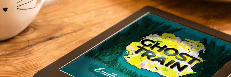 A close-up of a tablet or book-like device lying on a wooden table, displaying the title 'GHOST TRAIN' in large yellow letters over a dark illustrated background. A white ceramic teapot is partially visible at the upper left.
