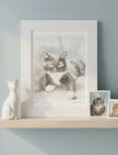 A framed monochrome cat sketch print displayed on a light wood shelf against a pale blue wall, with a white cat figurine on the left and two smaller framed cat photos on the right.