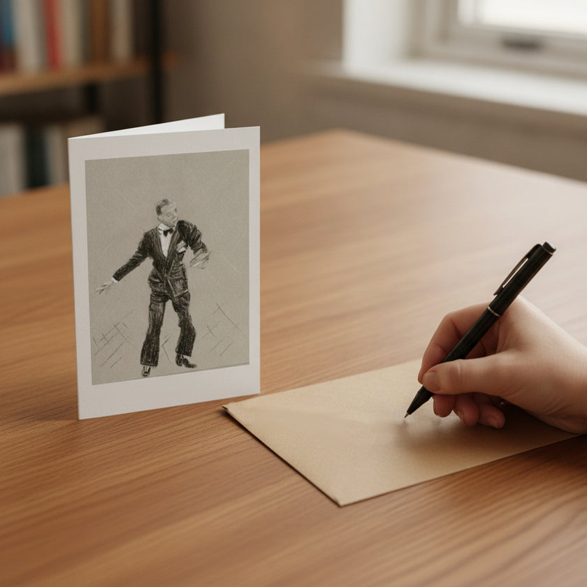 A hand holding a black pen writes on a beige card on a wooden desk, with a matching printed card featuring a black-and-white sketch of Fred Astaire standing nearby.