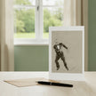 A greetings card featuring Fred astaire, displayed on a light wood table next to a gold envelope and a black pen, with a blurred window and green garden in the background.