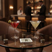 Two elegantly styled cocktail martinis on a glass table in a dimly lit bar setting. The left cocktail is a vibrant red martini with a lemon twist, and the right is a creamy yellow martini with a sugar rim. Fred Astaire Coaster lies on the table between the glasses