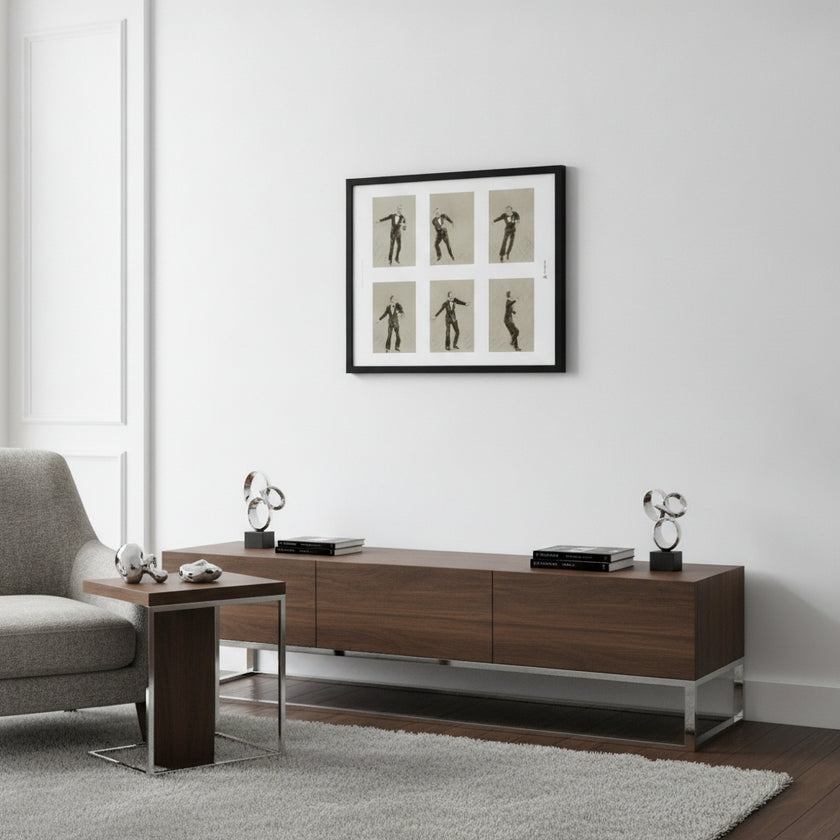 A modern living room scene featuring a dark brown wooden media console with silver metal legs, styled with decorative objects. Above it, a black-framed art print depicting six black-and-beige dance or gymnastic poses hangs on a white wall.