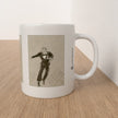 A white ceramic mug with a printed illustration of a man in a suit dancing, placed on a light wood surface against a white wall.