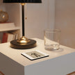 A modern black table lamp with a gold-toned base and pull chain sits on a white side table next to a clear glass tumbler and a small square photo card in a softly lit interior setting.