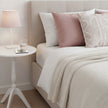 A styled bedroom scene showing a bed with white bedding, a dusty pink pillow, a textured cream throw blanket, and a beige upholstered headboard. A white side table with a pink lamp and a glass of water sits beside the bed.