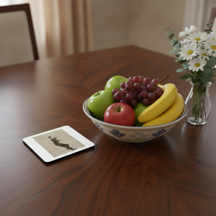 A ceramic fruit bowl filled with apples, grapes, and bananas sits on a polished dark wood dining table, next to a small vase of white daisies and a square coaster with a black-and-white photo print.