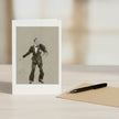 A greeting card with a black and white sketch of Fred Astaire, displayed on a light wooden table next to a brown envelope and a black pen.