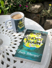 'Ghost Train' paperback by Emily Tellwright lies on a white outdoor patio table next to a matching white mug with the same cover design. The setting includes garden plants and rocks in the background.