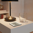 A square coaster with a black and white graphic print sits on a white side table next to a clear drinking glass and a black and gold table lamp.