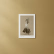 A framed art print featuring a sepia-toned sketch of a dancer in a long dress, displayed against a warm beige wall.