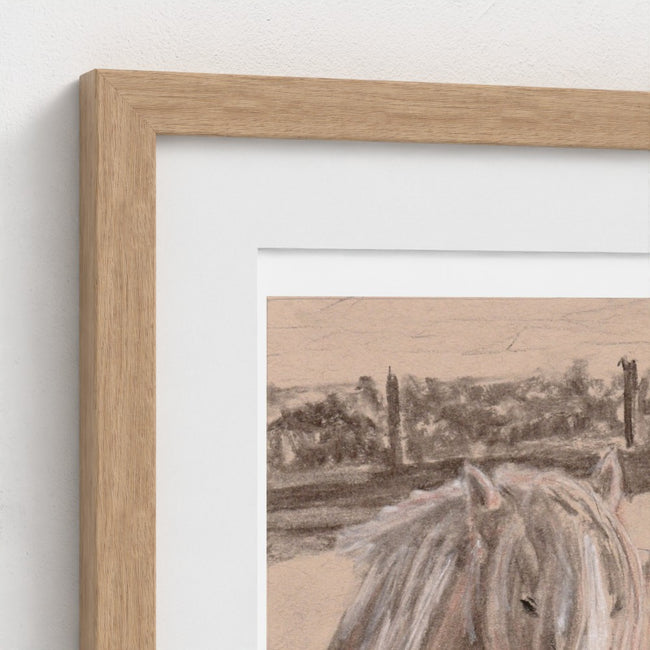 A close-up of a framed art print featuring a sepia-toned landscape with a horse in the foreground, set in a light oak frame with a white mount against a textured off-white wall.