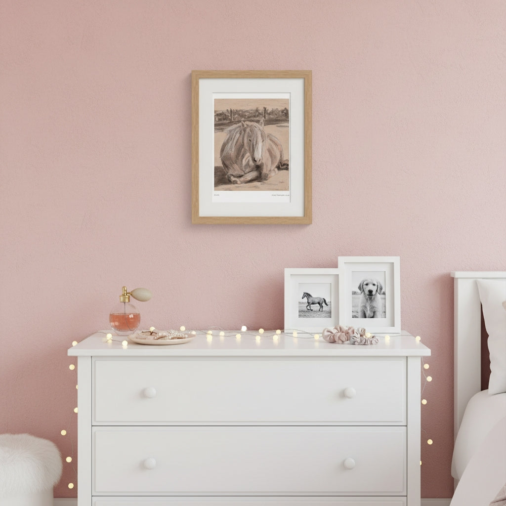 A framed sepia-toned horse print hangs on a dusty pink wall above a white dresser in a styled bedroom. Two smaller framed black-and-white horse prints sit on the dresser, with warm string lights draped around it.