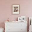 A framed sepia-toned horse print hangs on a dusty pink wall above a white dresser in a styled bedroom. Two smaller framed black-and-white horse prints sit on the dresser, with warm string lights draped around it.