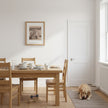 A framed art print of a drawing of a pony is on the wall above a light oak dining table with matching wooden chairs in a bright, modern dining room. A golden retriever dog is lying on a grey rug on the floor next to the table.