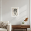 A black and white sketch print of a stylized face hangs on a light gray wall in a modern living room setting above a wooden console table with a vase and books.