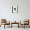 A framed black and white sketch portrait of a person hangs on a white wall above a mid-century modern wooden coffee table, flanked by two matching armchairs with beige cushions. A bottle of red wine and two wine glasses sit on the table.