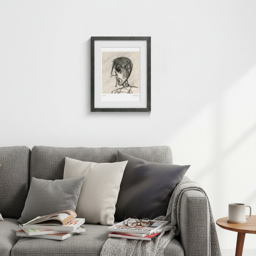 A framed black and white sketch portrait of a person hangs on a white wall above a gray sofa with pillows, books, and a throw blanket in a bright living room.