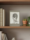 A framed pencil portrait drawing of a person in profile, displayed on a wooden bookshelf alongside books and a potted plant.