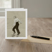A vertical art print card featuring a black and white drawing of ginger rogers, displayed on a light wood tabletop next to a black pen and a beige envelope.