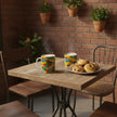 Two colorful patterned mugs sit on a wooden table in a cafe setting, with a plate of cookies beside them and a brick wall with hanging plants in the background.